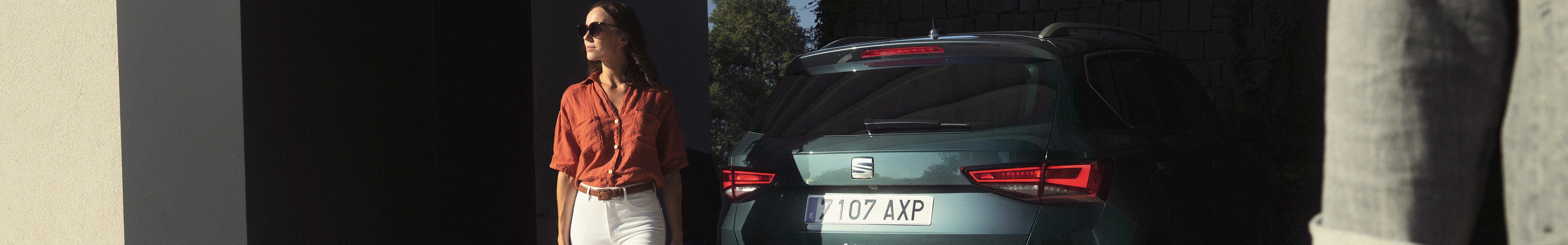 A photo of a woman about to enter her car, she is looking at the camera. The woman is wearing an ecru coloured blazer, and has brown curly hair.